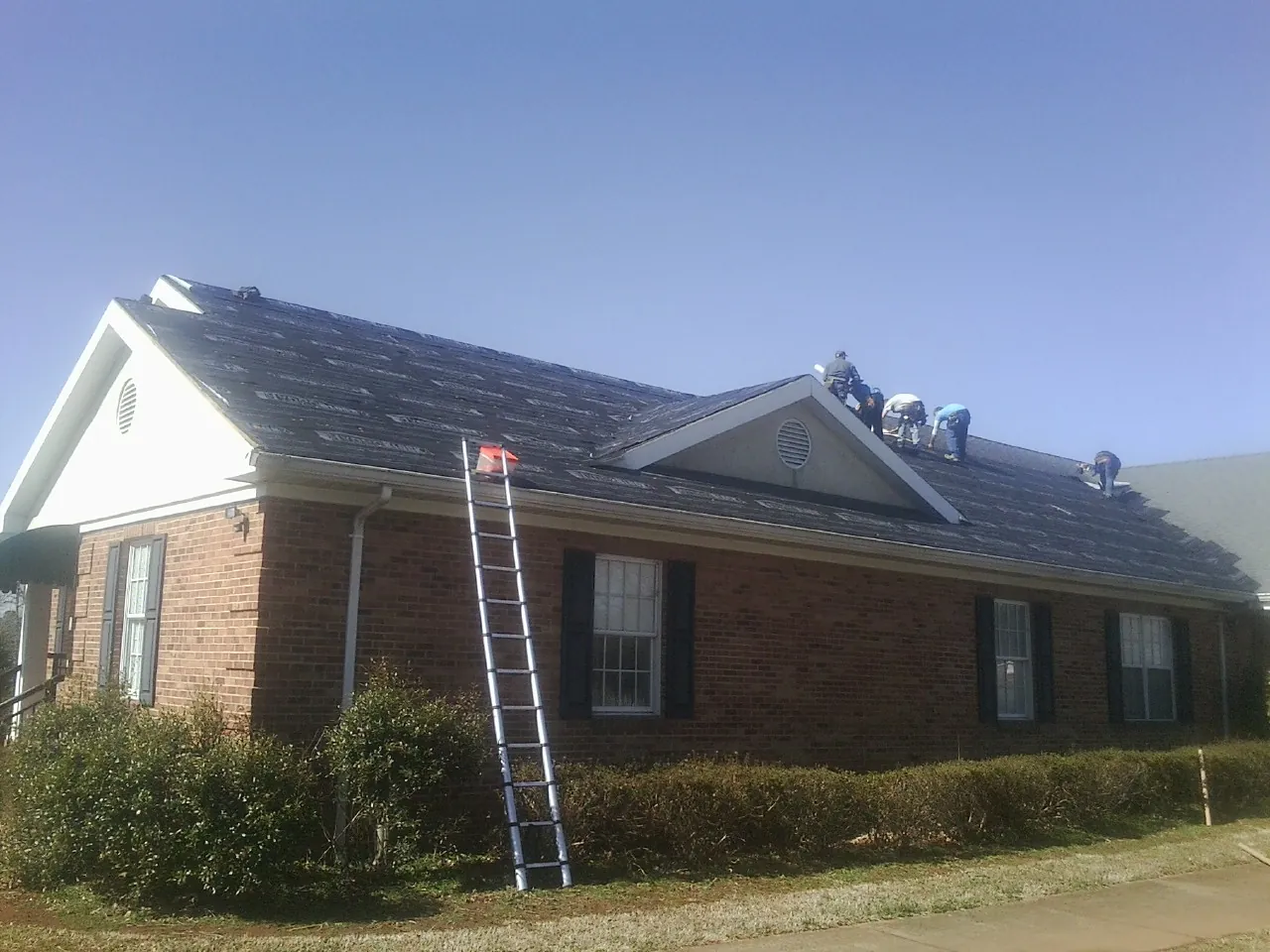 Crew tearing shingles off a brick ranch roof with a ladder set — Emergency Roof Repair work by our crew in White Lake
