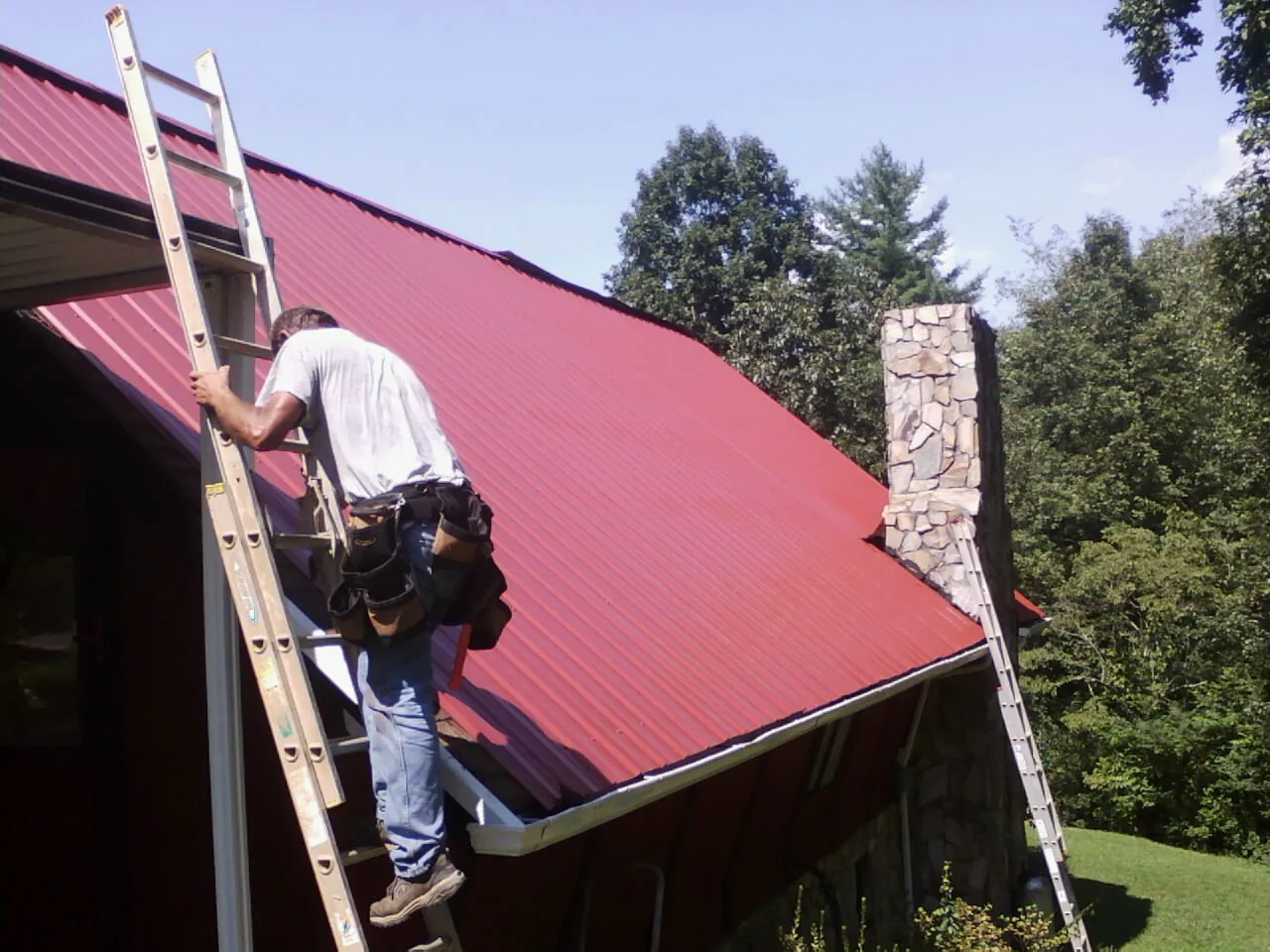 Roofer on a ladder beside a red metal roof and stone chimney — Soffit & Fascia Repair by Roofing Brothers in White Lake